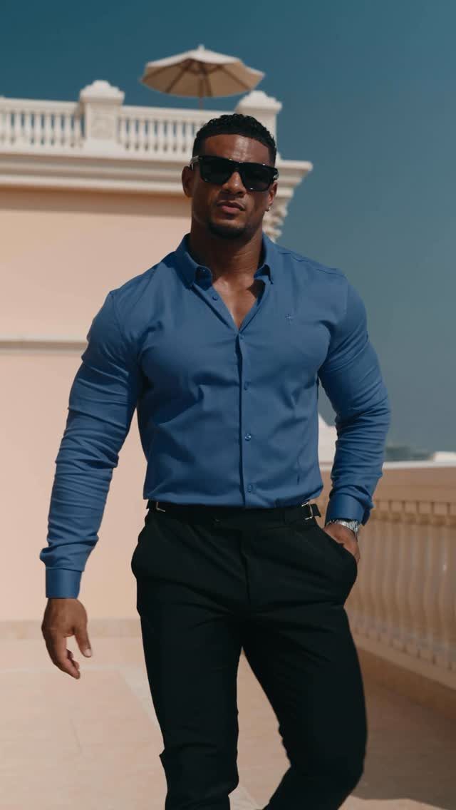 A muscular man in a blue shirt and black trousers posing on a sunny balcony.