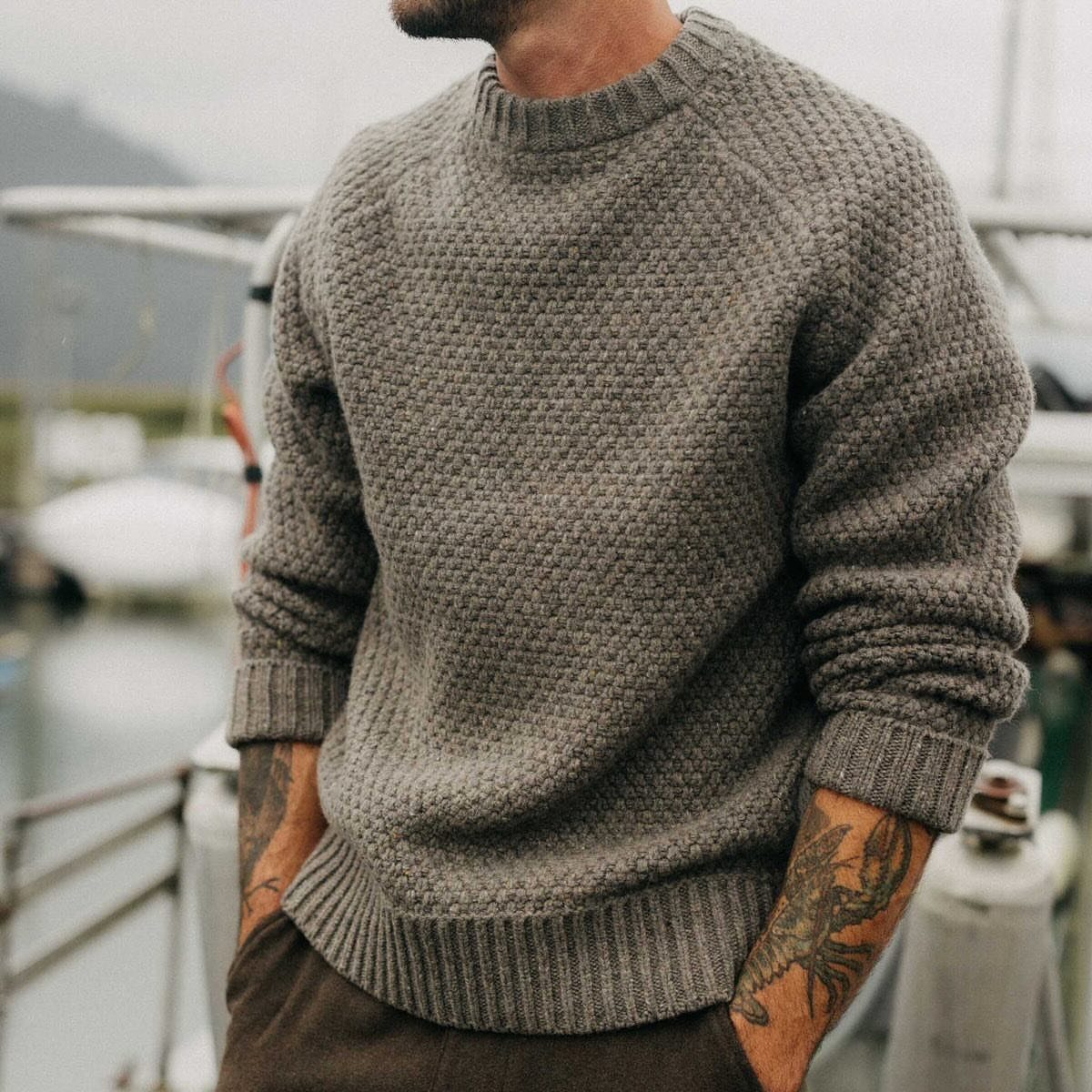 Man wearing a textured grey crewneck knit sweater and brown trousers standing near a boat.