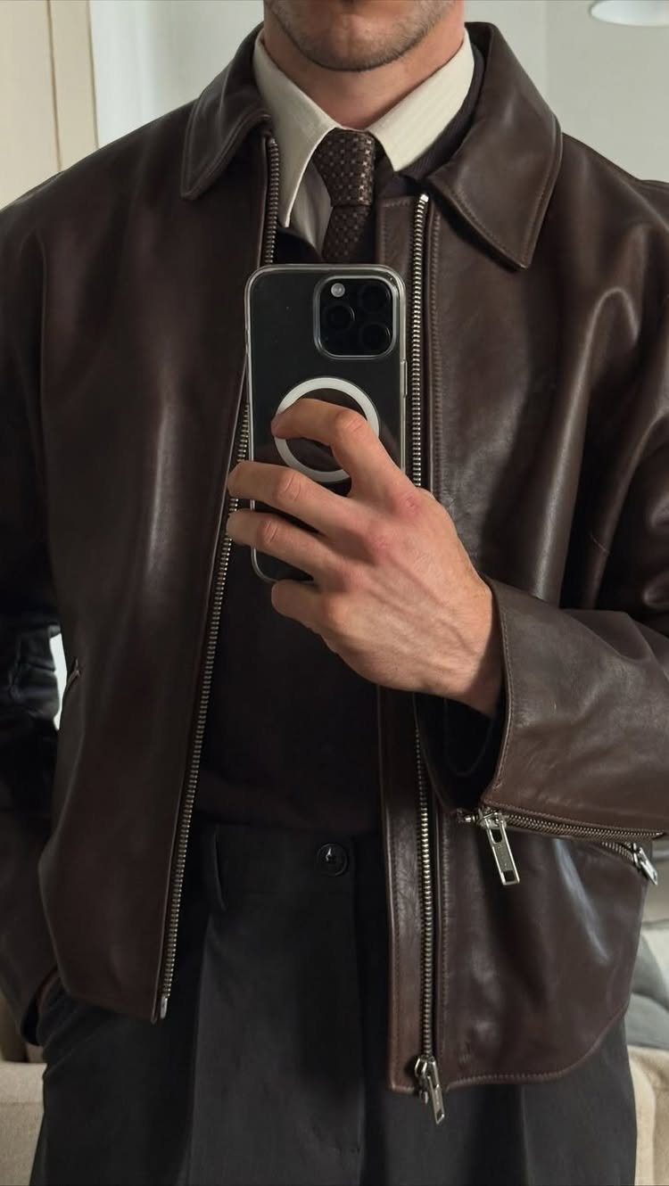 Man in a brown leather jacket, white shirt, and patterned tie taking a mirror selfie.