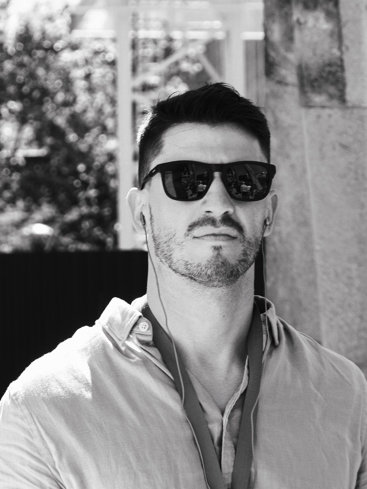 Man with stubble wearing dark minimalist sunglasses and a light button-up shirt.