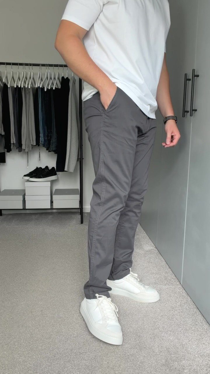A man wearing a white t-shirt, grey chinos, and white sneakers standing indoors.