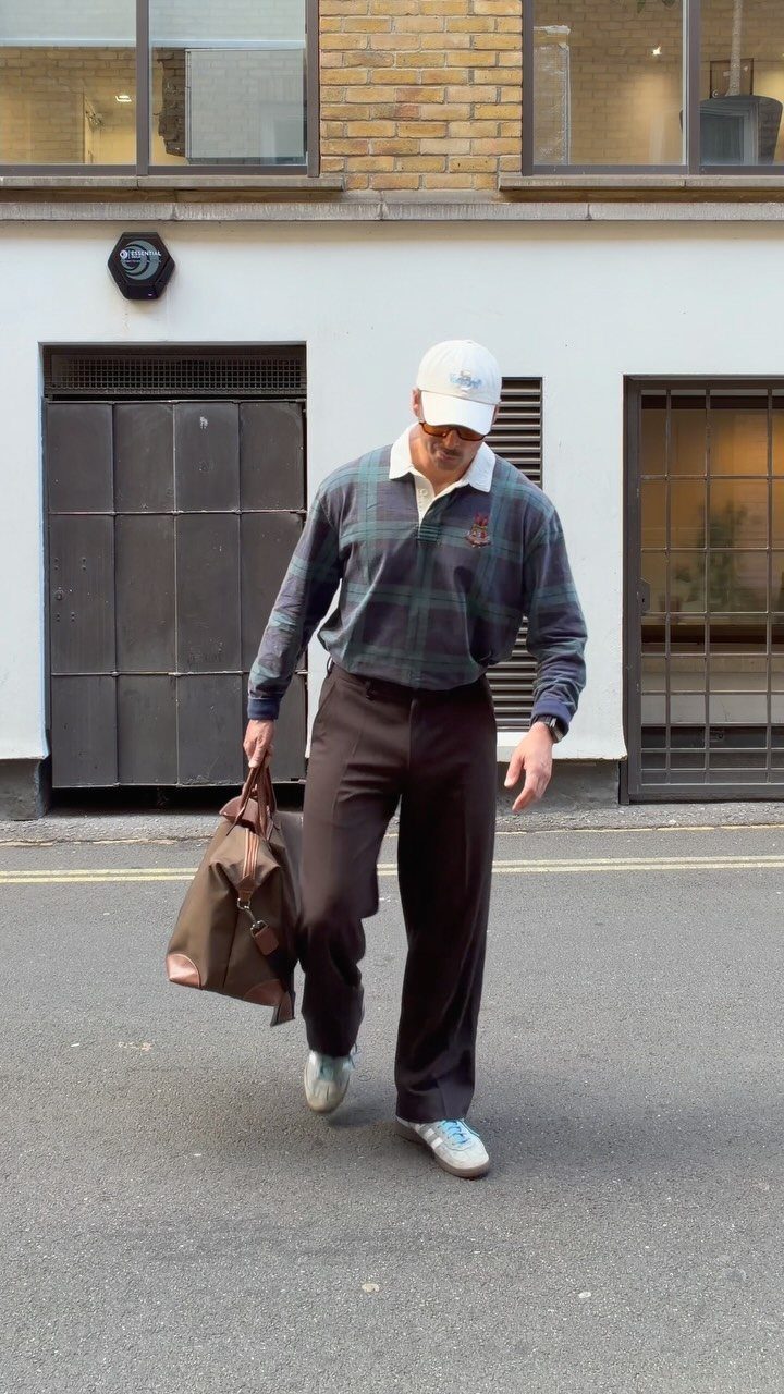 Middle-aged man in plaid polo and brown trousers walking through a city street carrying a leather bag.