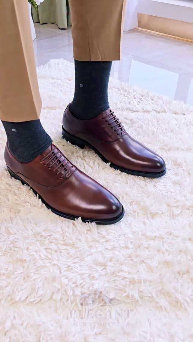 Polished dark brown leather derby shoes worn with grey socks and tan trousers on a white rug.