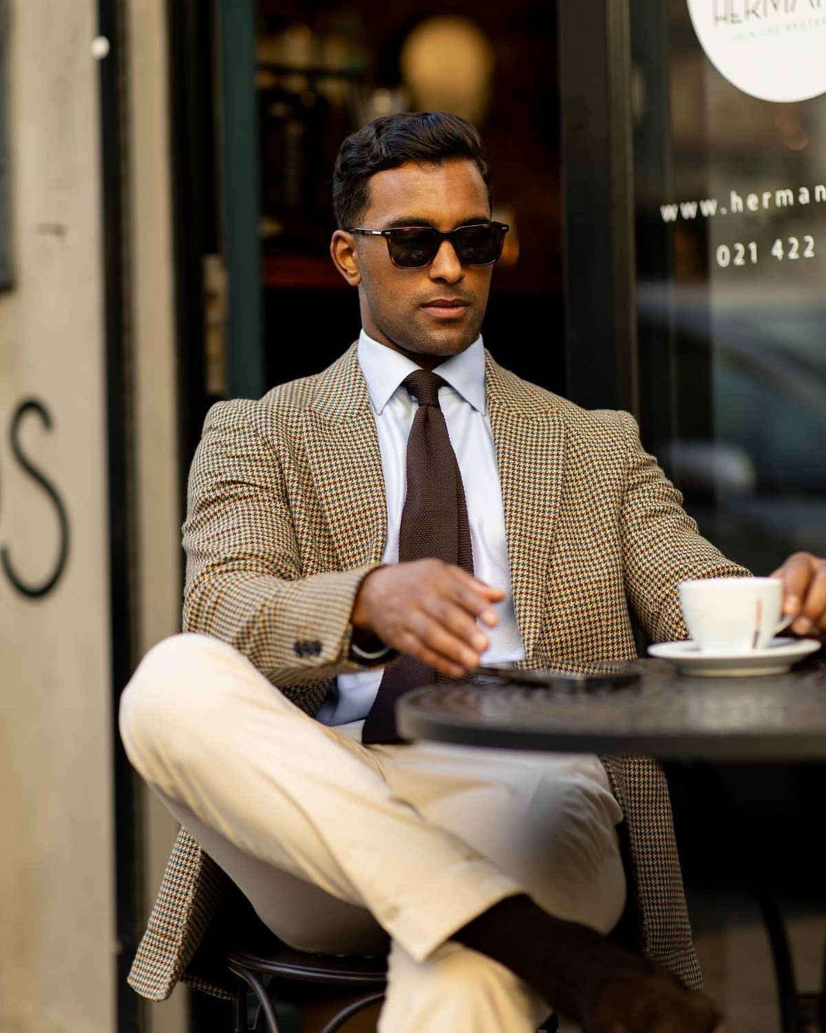 Man in patterned blazer, blue shirt, and tie sitting at an outdoor cafe table.