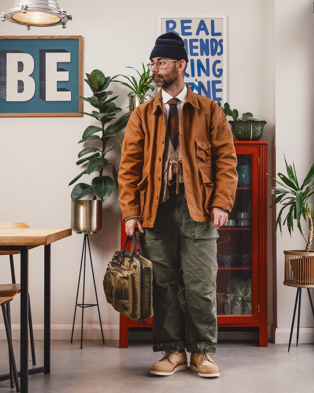 Man in his 40s wearing a brown utility jacket, tie, and olive cargo pants standing indoors.