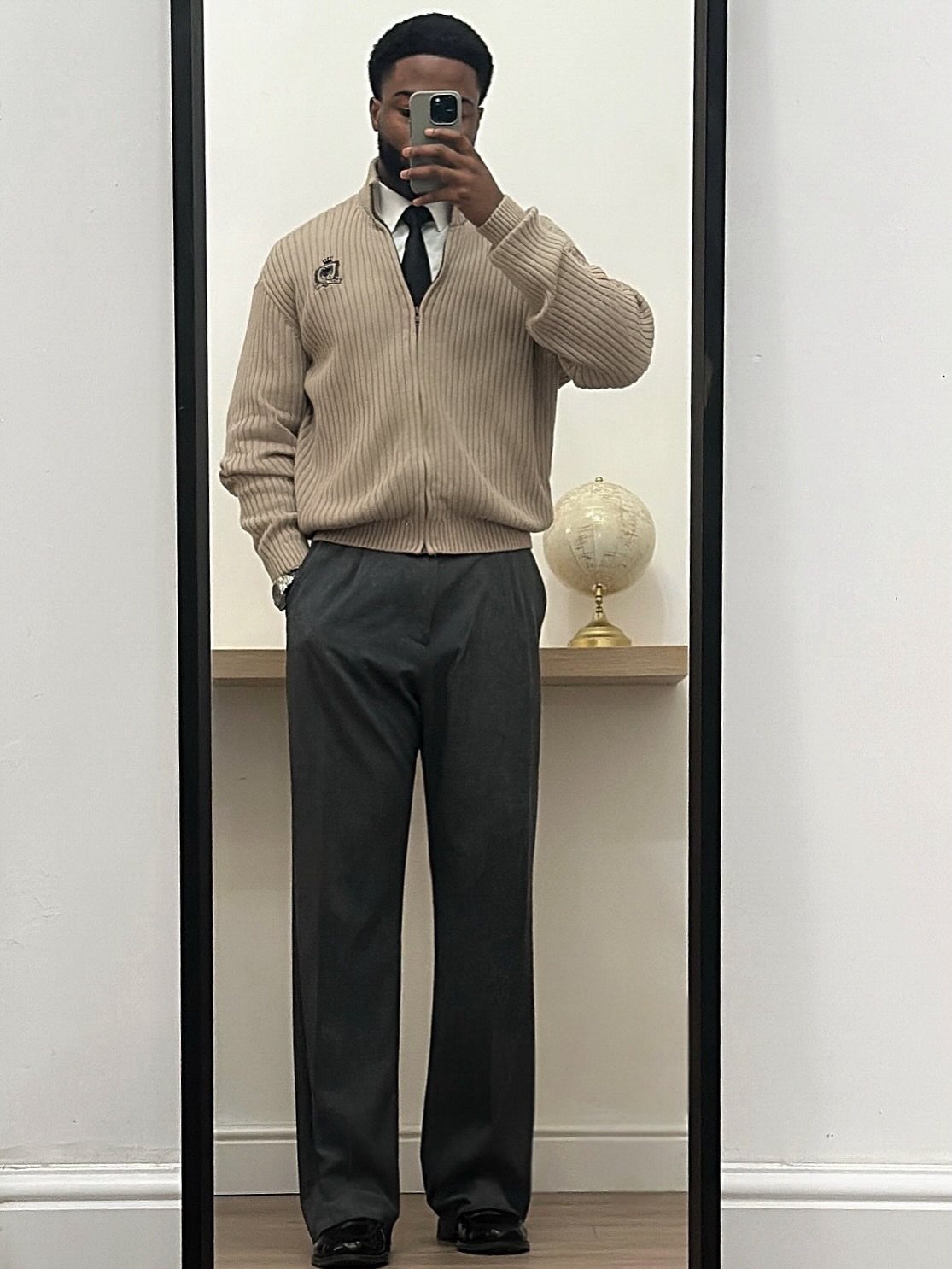 Man in a beige cardigan, white shirt, black tie, and grey trousers posing for a mirror selfie.