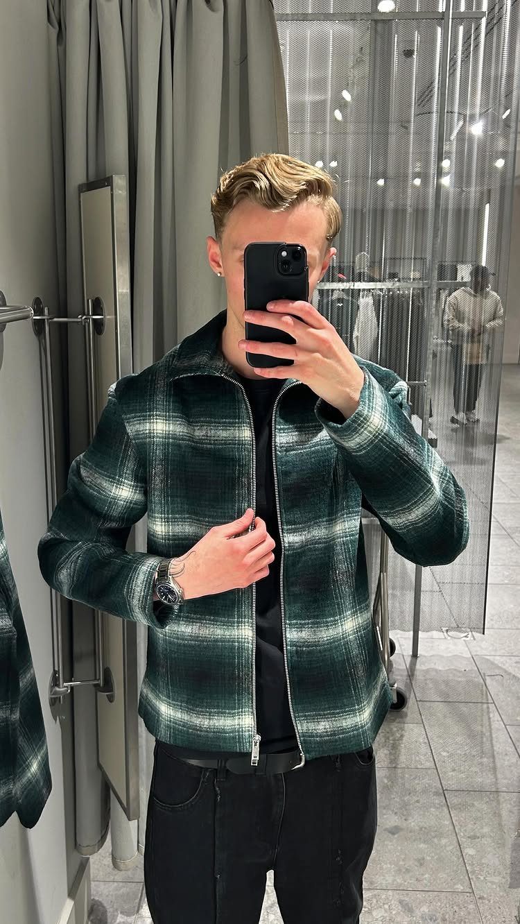 Man in a green plaid flannel jacket and black pants taking a mirror selfie.