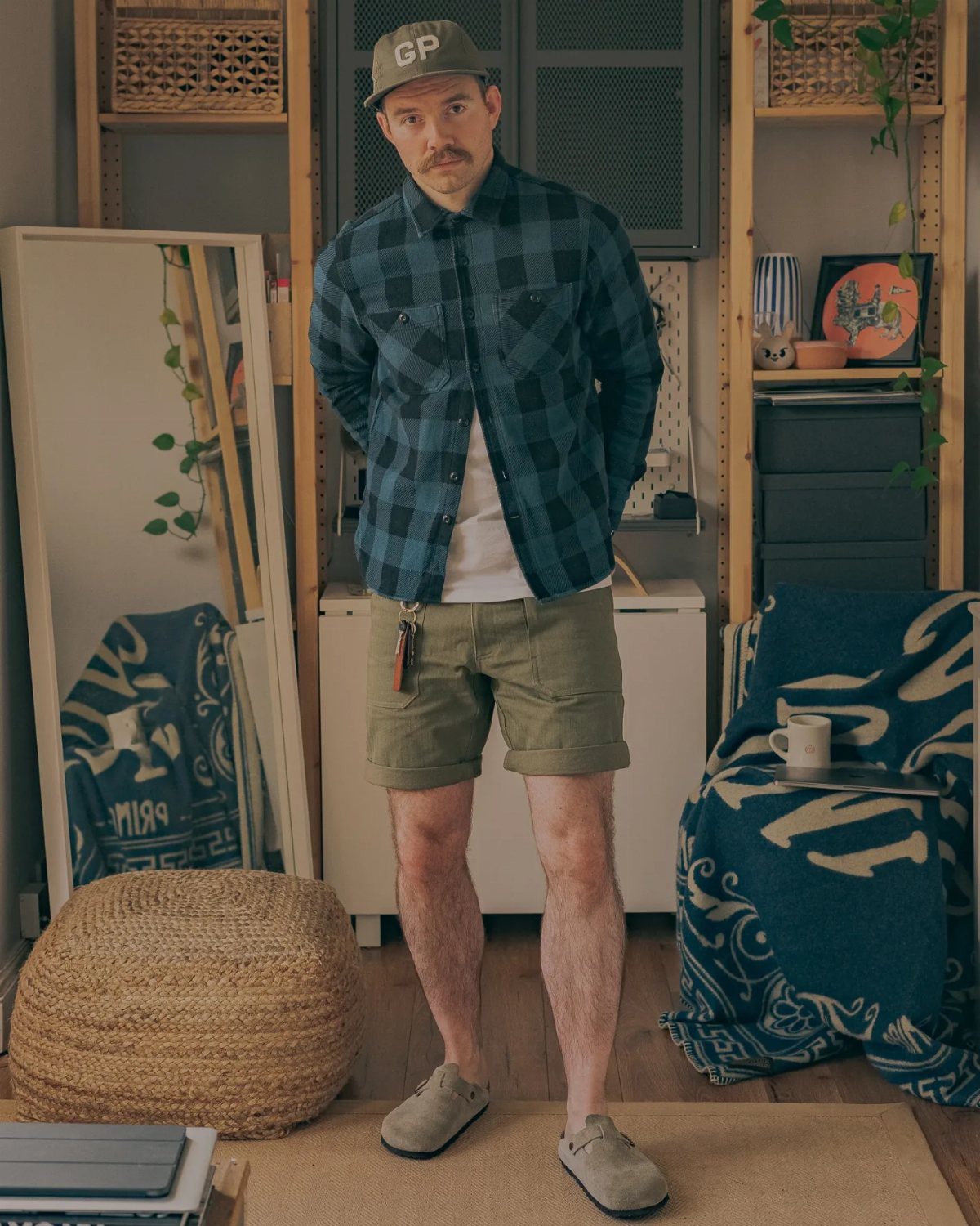 Man wearing a blue plaid flannel shirt, white t-shirt, olive shorts, and grey shoes.