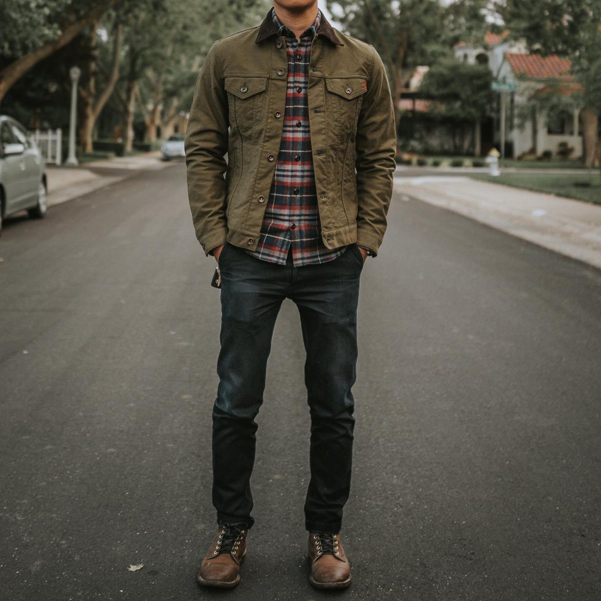 Man wearing a plaid flannel shirt, olive jacket, dark jeans, and brown boots on a street.