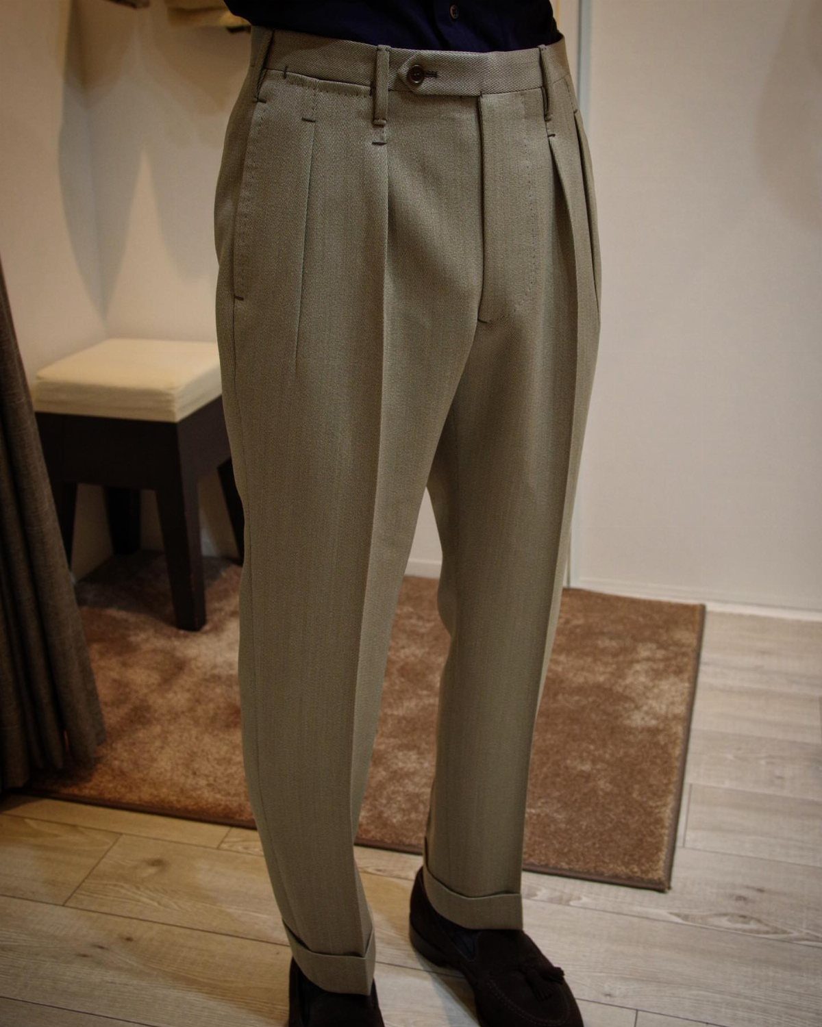 Man wearing light beige pleated trousers and dark loafers standing on a wooden floor.