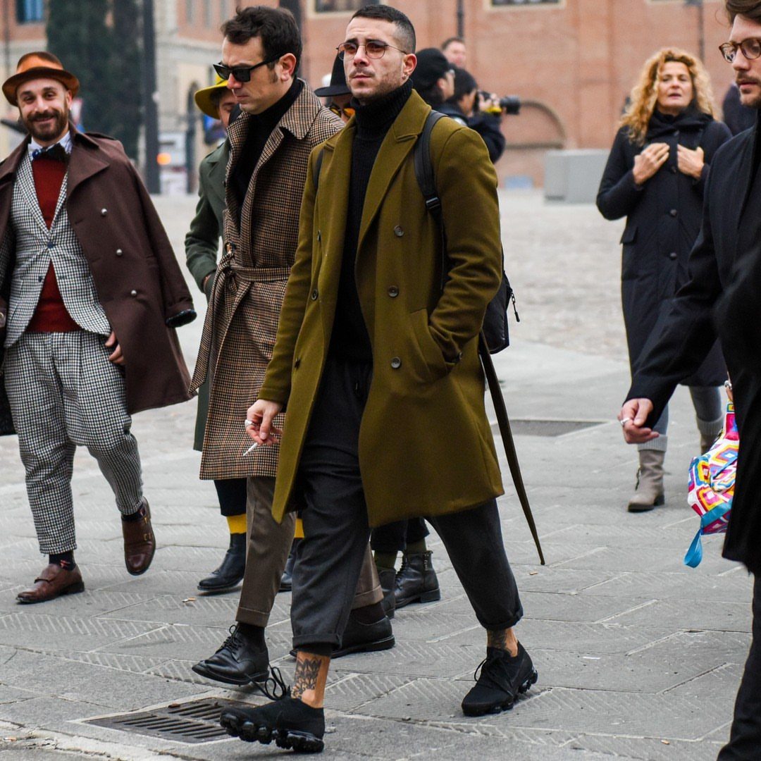 Man walking in a city wearing an olive green coat and black cropped trousers.