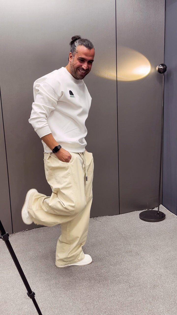 Man in white sweatshirt and cream drawstring pants posing in a grey studio.