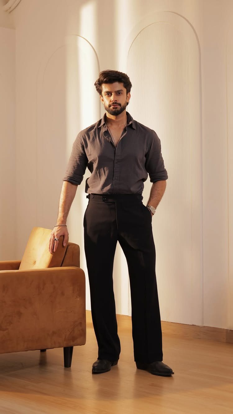 Man in a grey shirt and black dress pants posing next to a brown armchair.