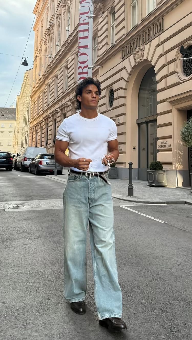 A man in a white t-shirt and light blue wide-leg jeans standing on a city street.