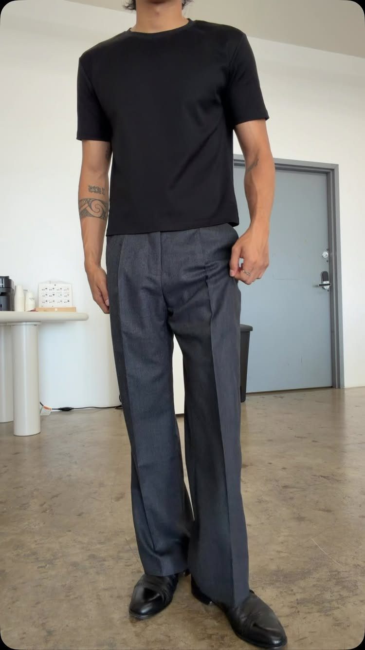 A man standing in a room wearing a black t-shirt and grey pleated trousers.