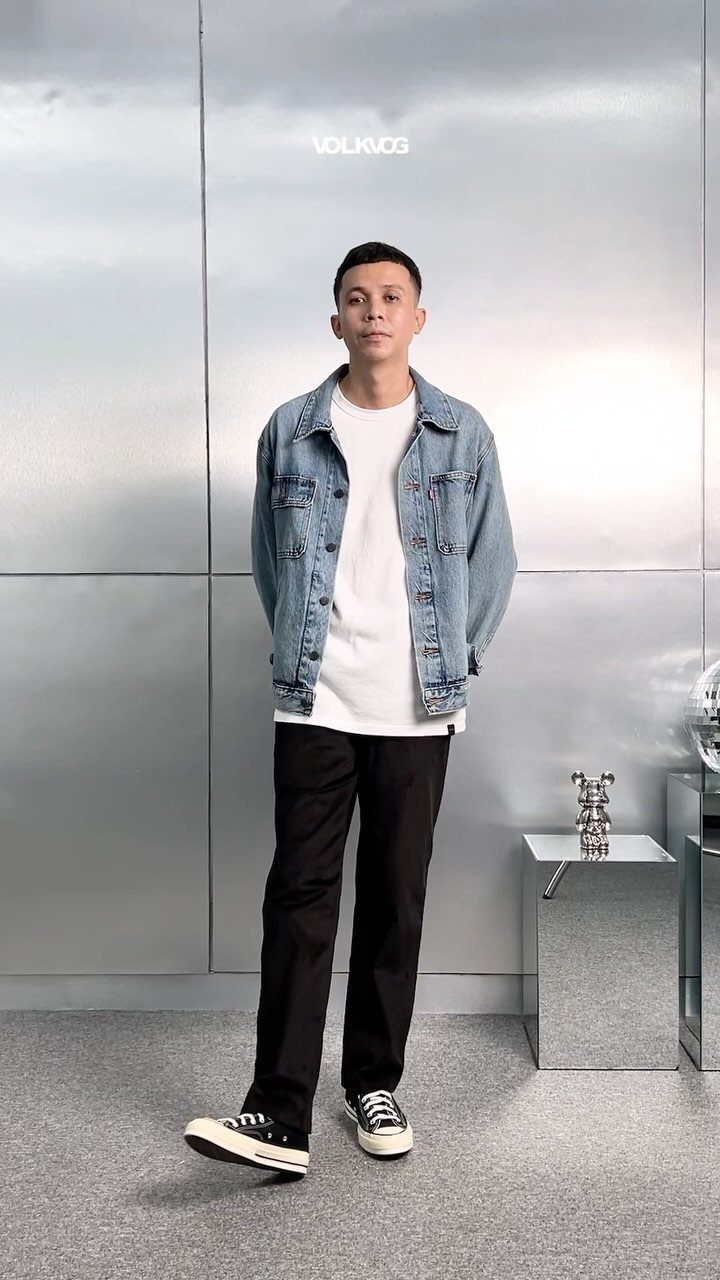 A man wearing a denim jacket, white t-shirt, and black pants standing against a silver wall.