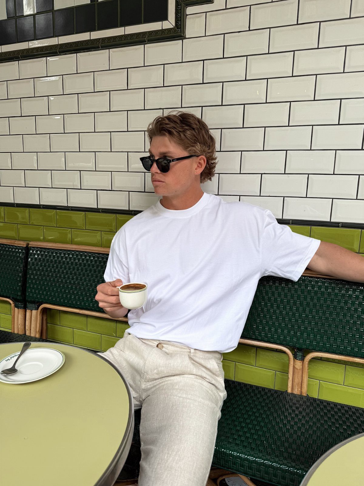 Man in white t-shirt and beige linen trousers sitting at a cafe table with coffee.