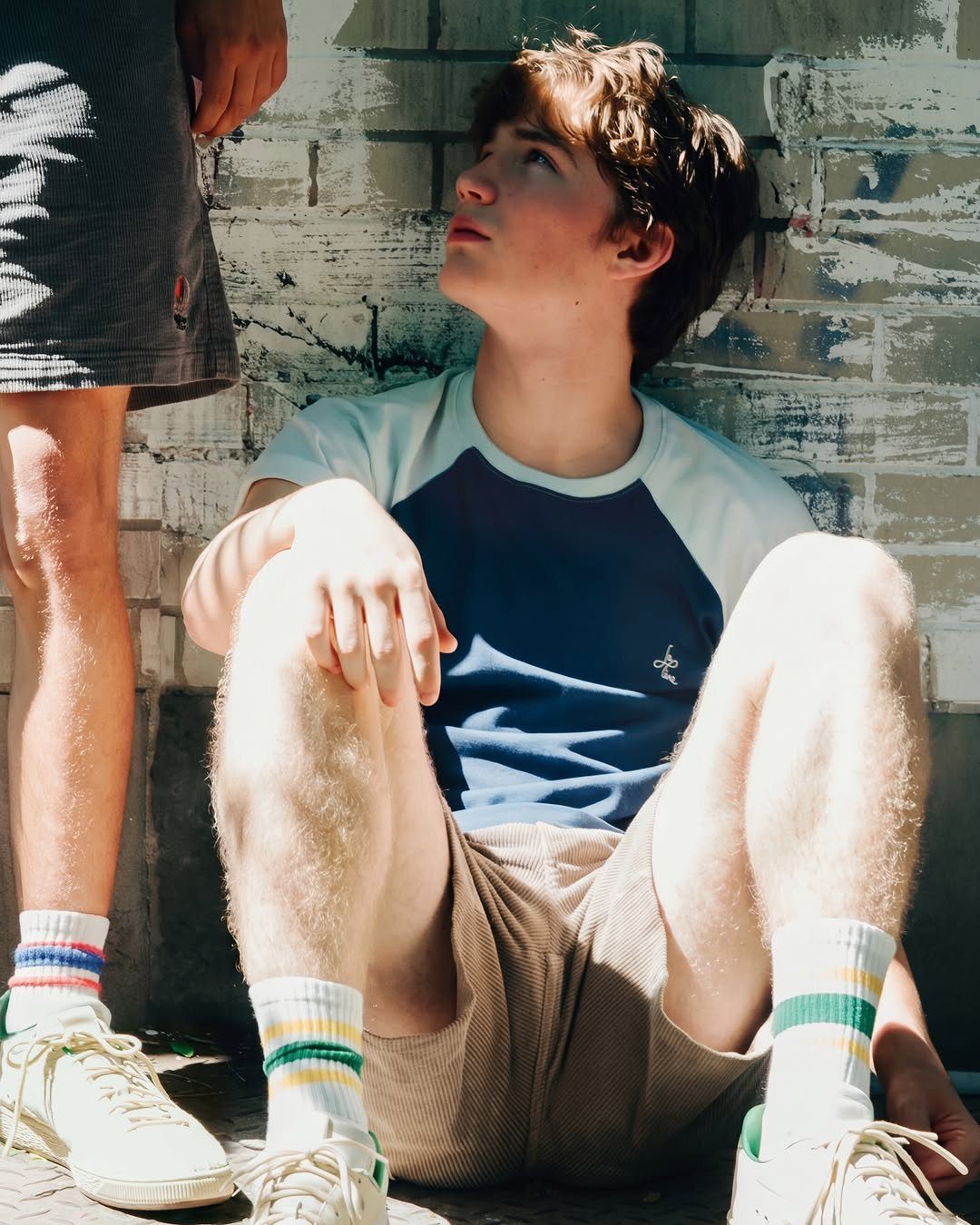 A young man in a blue raglan shirt and tan shorts sits against a white brick wall.
