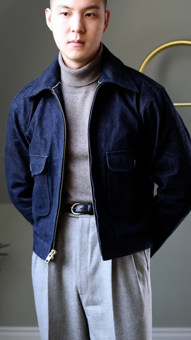 Man wearing a dark navy cropped worker jacket, grey turtleneck, and light grey pleated trousers.