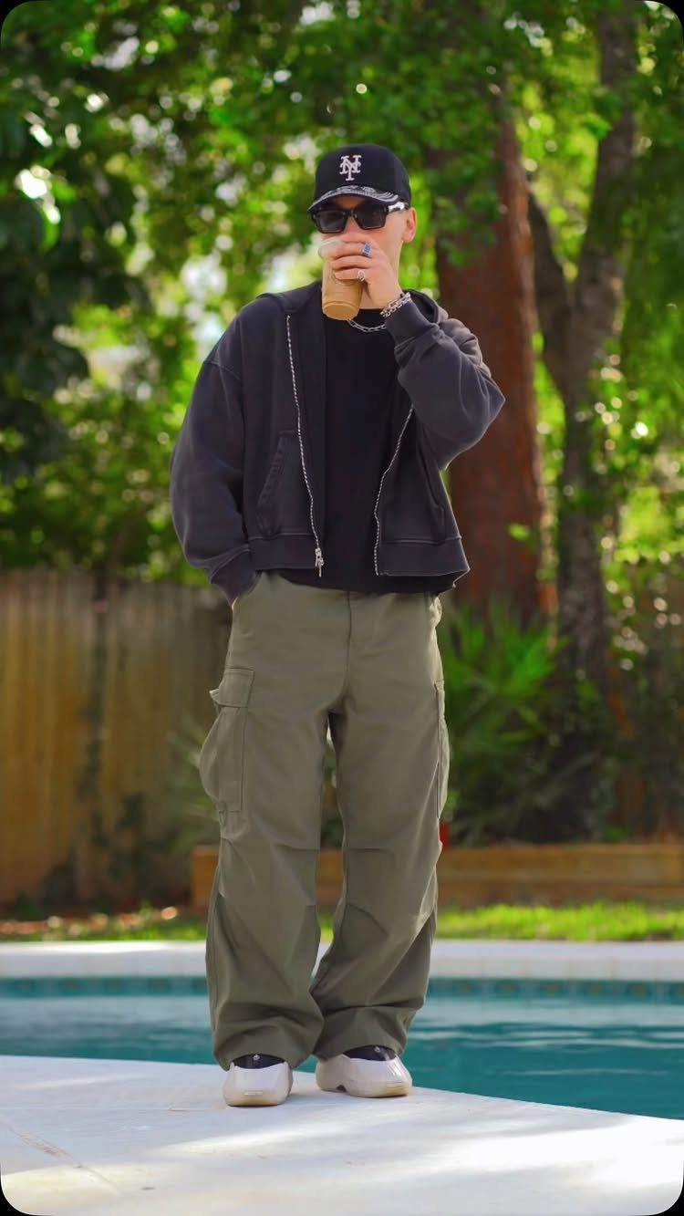 Man in olive cargo pants and dark jacket standing by a swimming pool.
