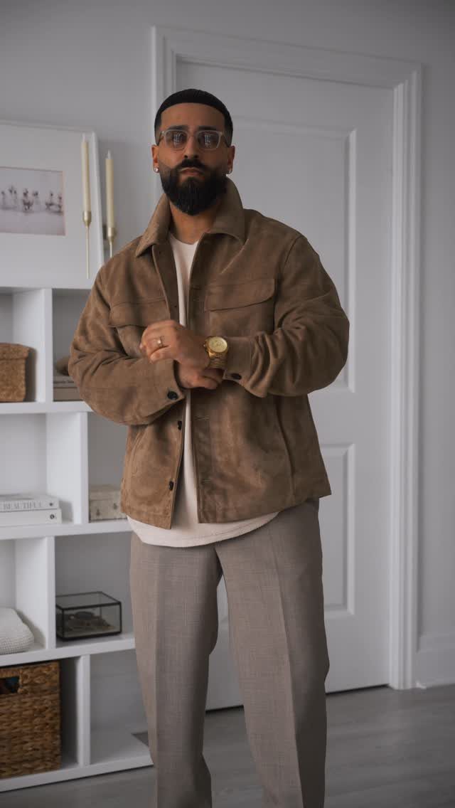 A man wearing a brown suede overshirt over a white t-shirt and grey trousers.