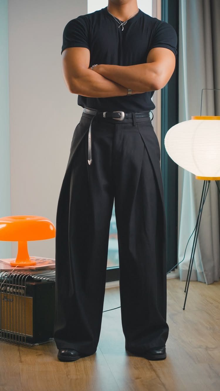 Man wearing black short-sleeved shirt and wide-leg black pleated trousers standing indoors.