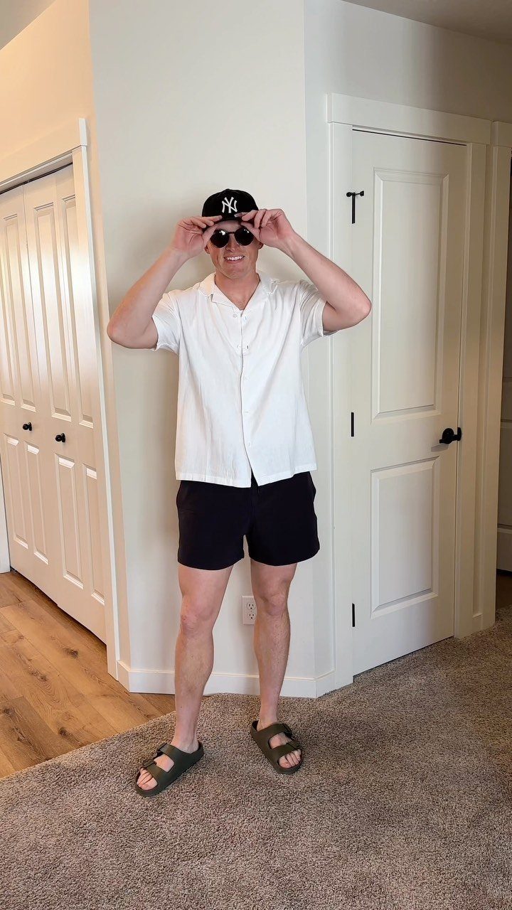 Man wearing a white camp collar shirt, black shorts, and a baseball cap standing indoors.