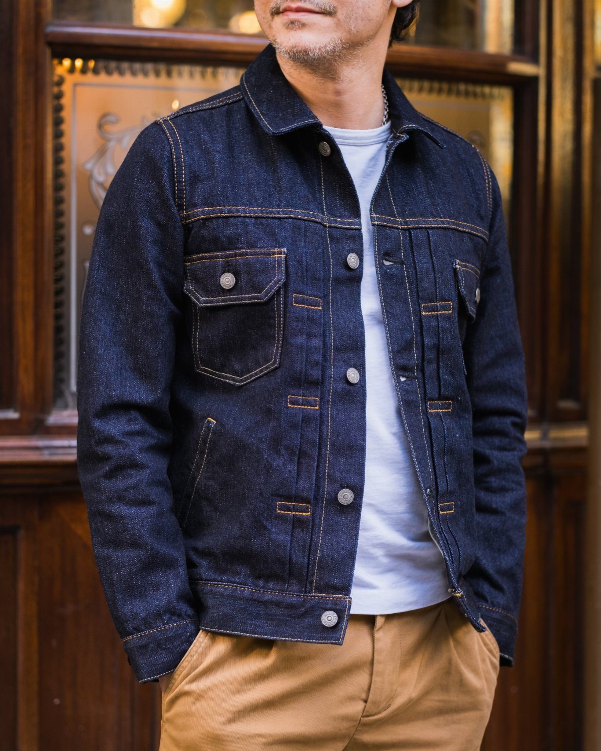 A man wearing a dark blue denim jacket, white t-shirt, and tan pants.