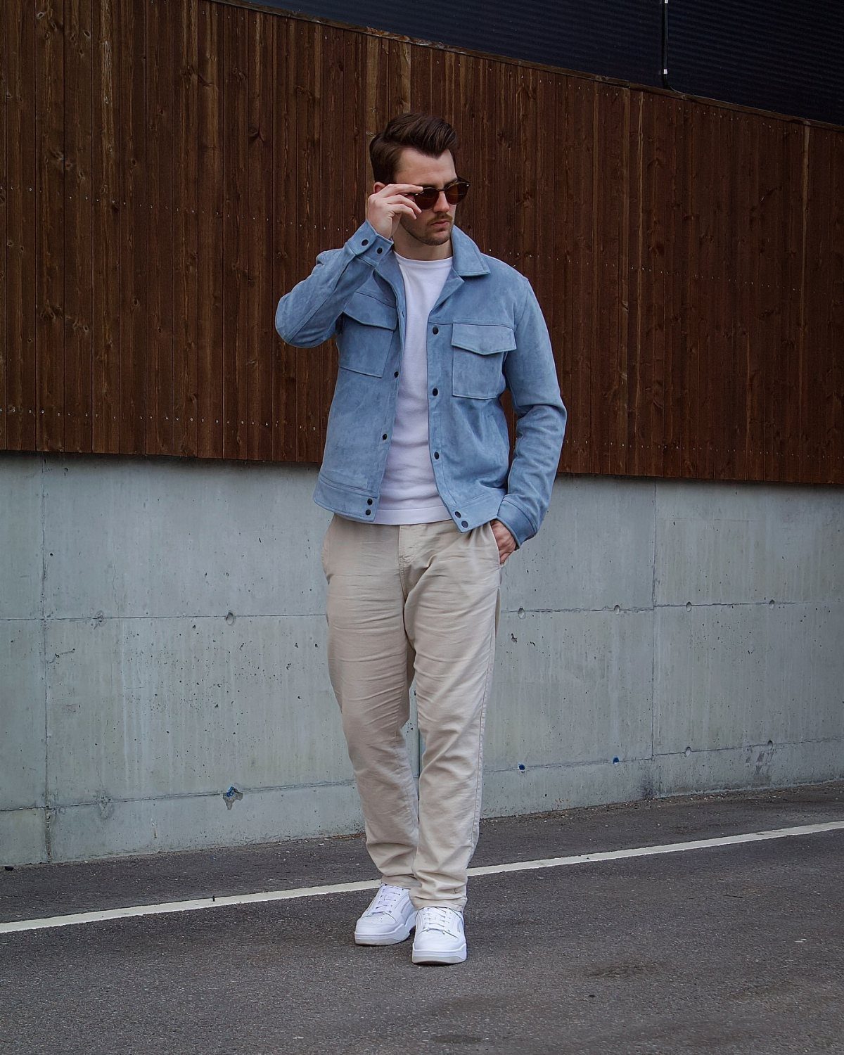 Man wearing a light blue jacket, white t-shirt, beige pants, and white sneakers standing against a wall.