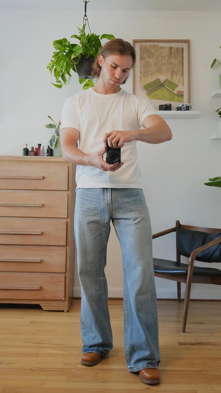 A man in a white t-shirt and flared blue jeans holding a small camera indoors.