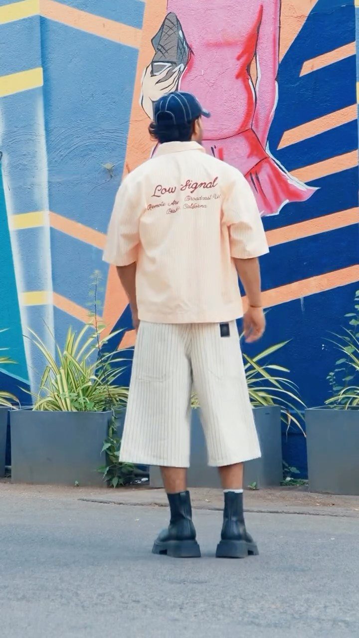 Man wearing cream short-sleeved shirt, wide-leg striped shorts, and black boots standing before a colorful mural.