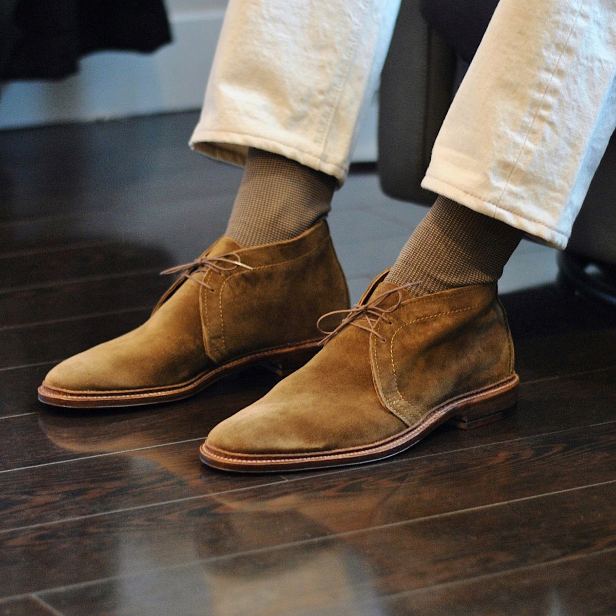 Tan suede chukka boots worn with brown socks and off-white trousers on a dark wood floor.