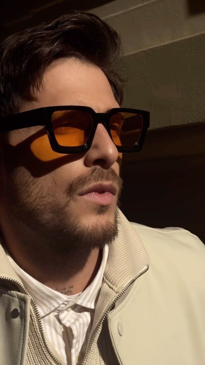 Man wearing black-framed sunglasses with amber tinted lenses and a cream jacket.