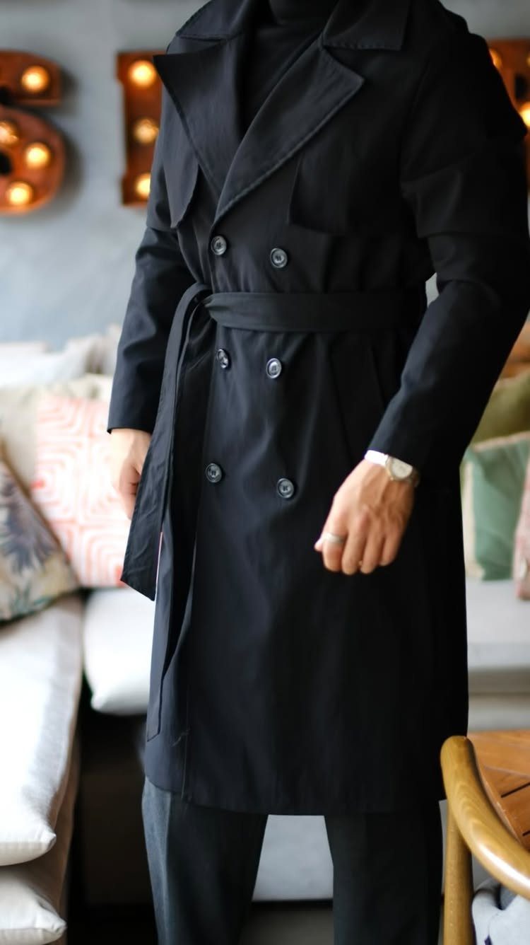 A man wearing a stylish black double-breasted trench coat with a waist belt.