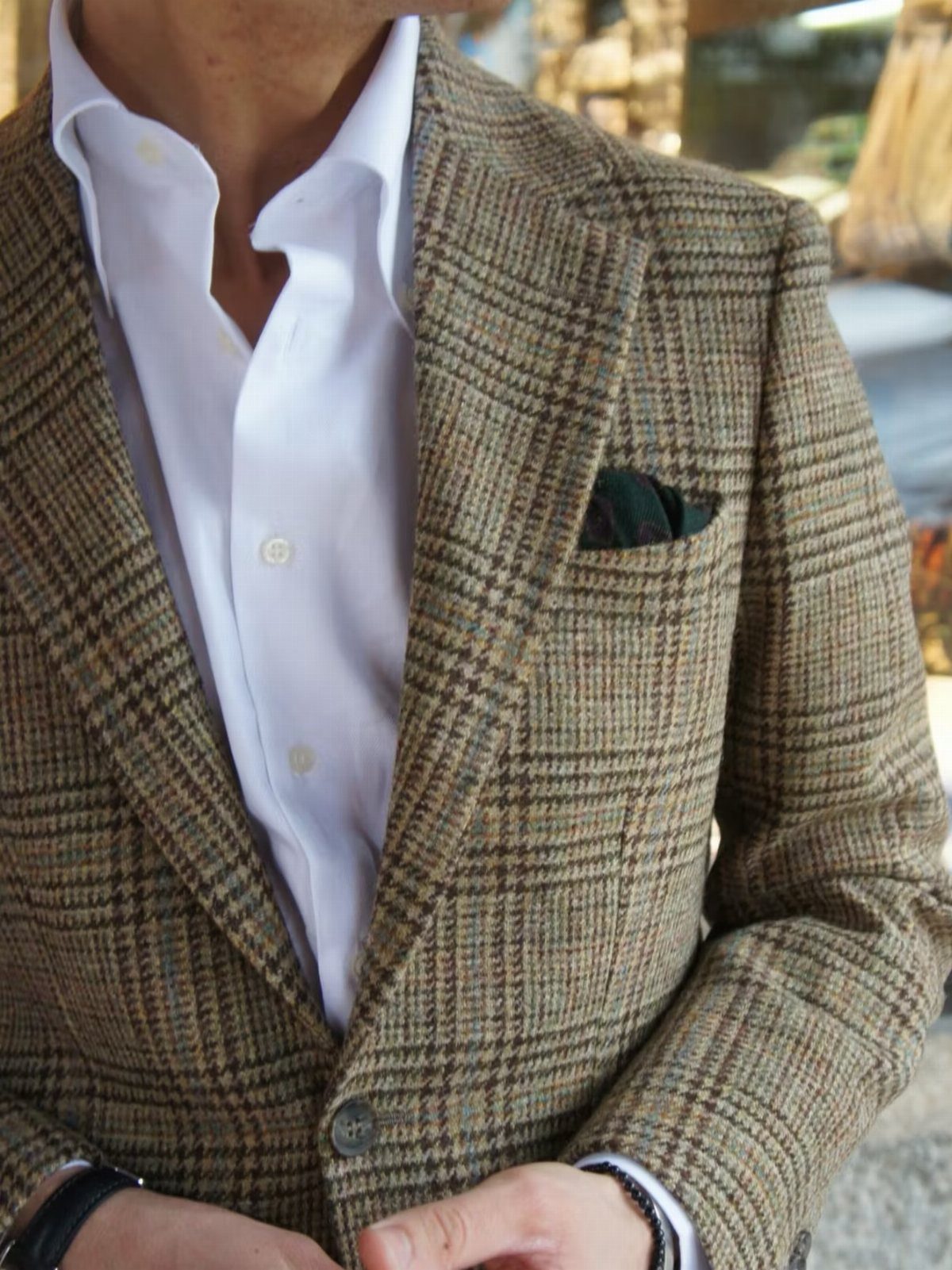 A man wearing a brown patterned tweed jacket over a white button-down shirt with a green pocket square.