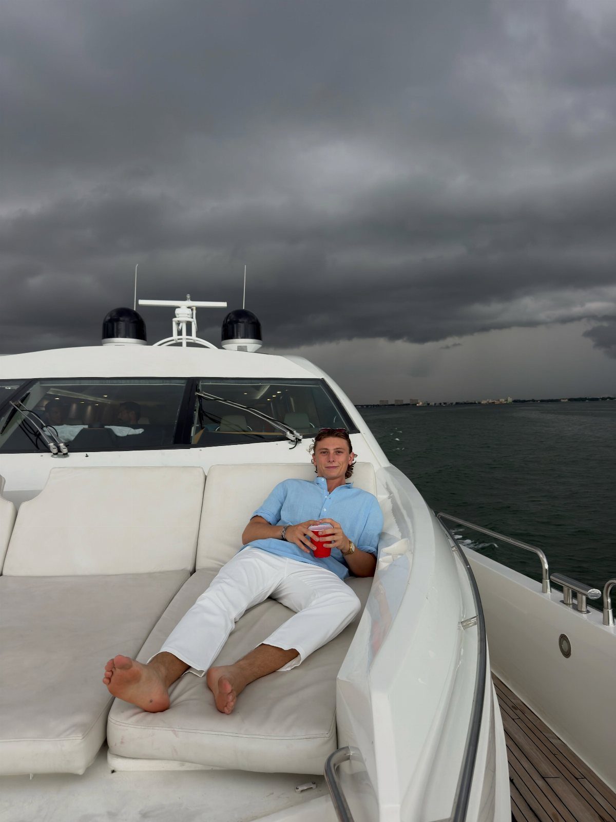 Man in light blue shirt and white pants relaxing on a white boat deck under stormy skies.