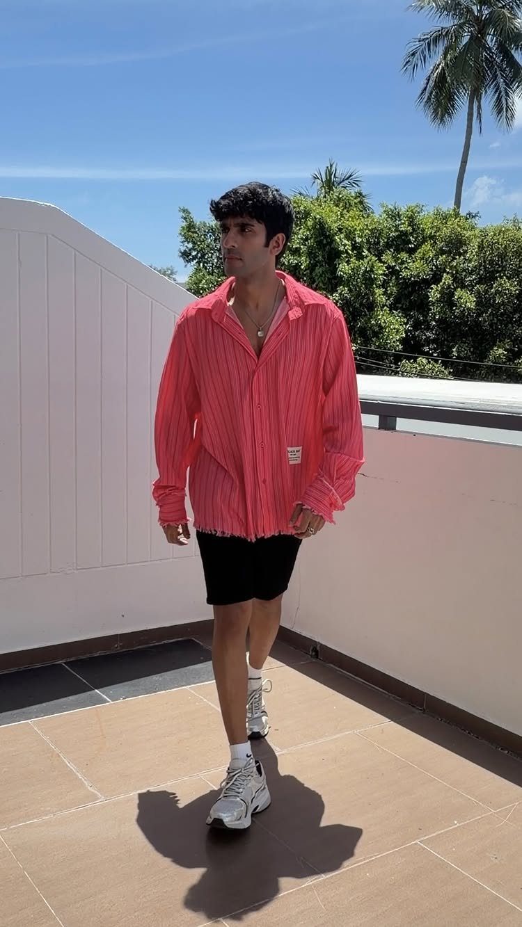 A man walking in a pink striped shirt, black shorts, and white sneakers under a blue sky.