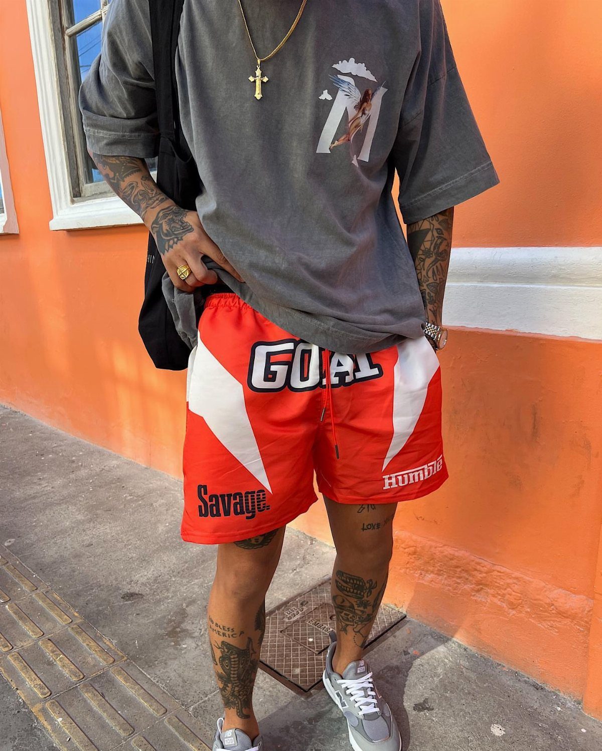 Man wearing a grey graphic t-shirt, red athletic shorts, and grey sneakers standing against an orange wall.