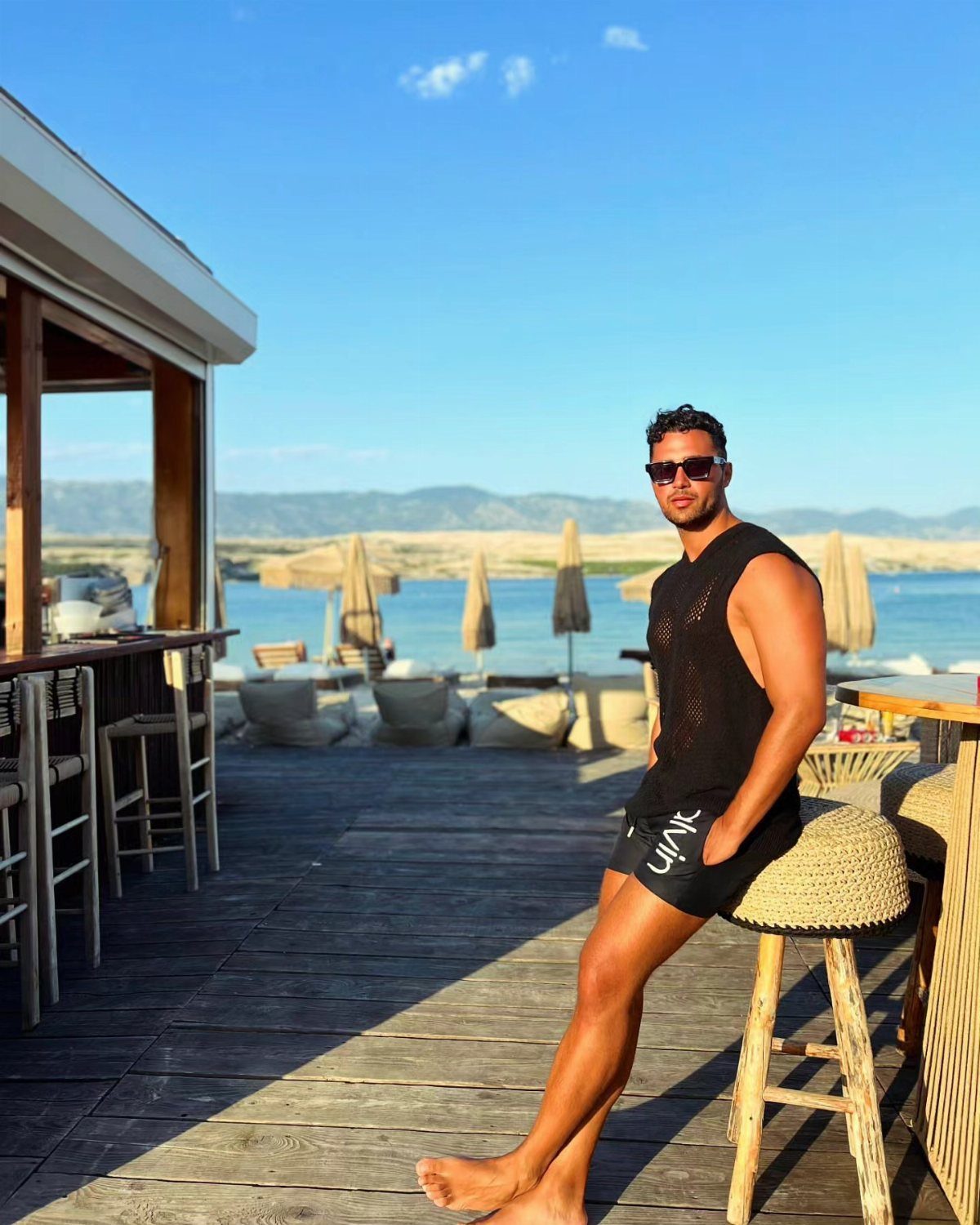 A man in a black knit tank top and swim trunks sitting on a stool at a beach club.
