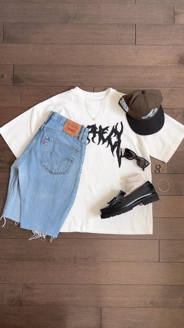 Black loafers, denim shorts, a white graphic t-shirt, and accessories laid out on a wooden floor.