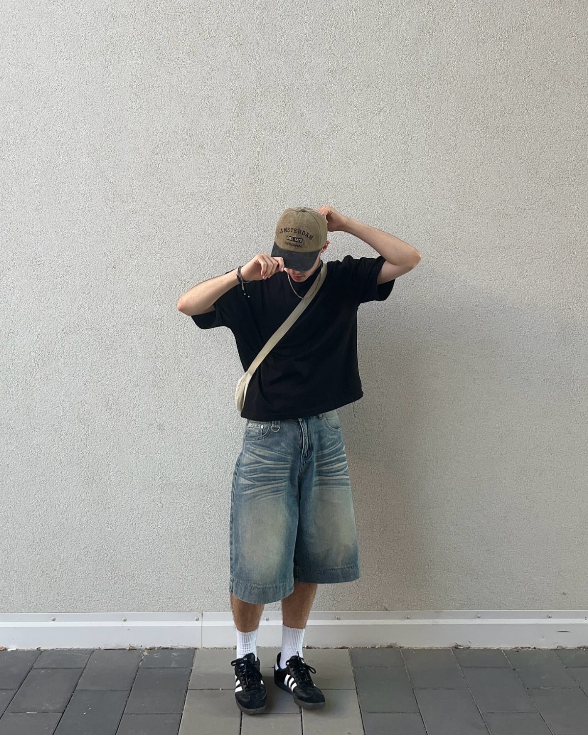 A person wearing a black t-shirt, baggy denim shorts, and a tan baseball cap against a grey wall.
