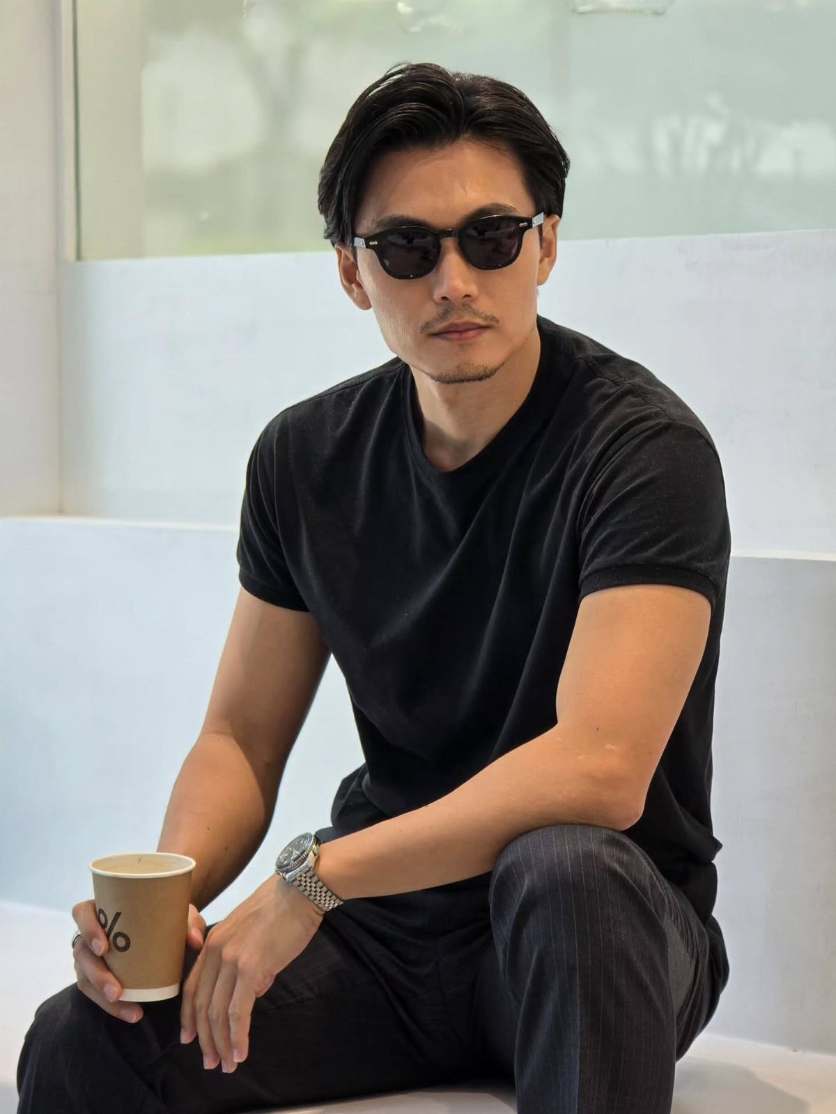 Man in a black t-shirt and dark sunglasses sitting and holding a coffee cup.