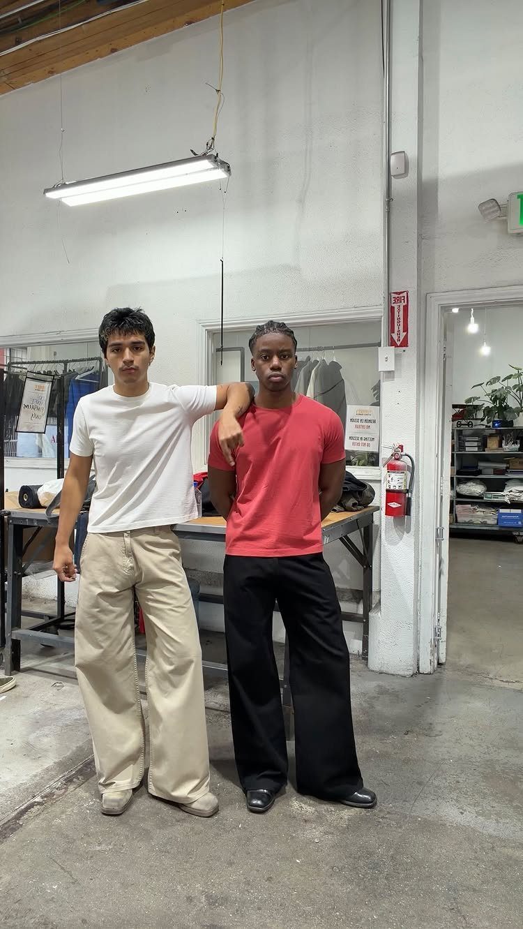 Two men posing in a workshop wearing white and red t-shirts with wide-leg trousers.