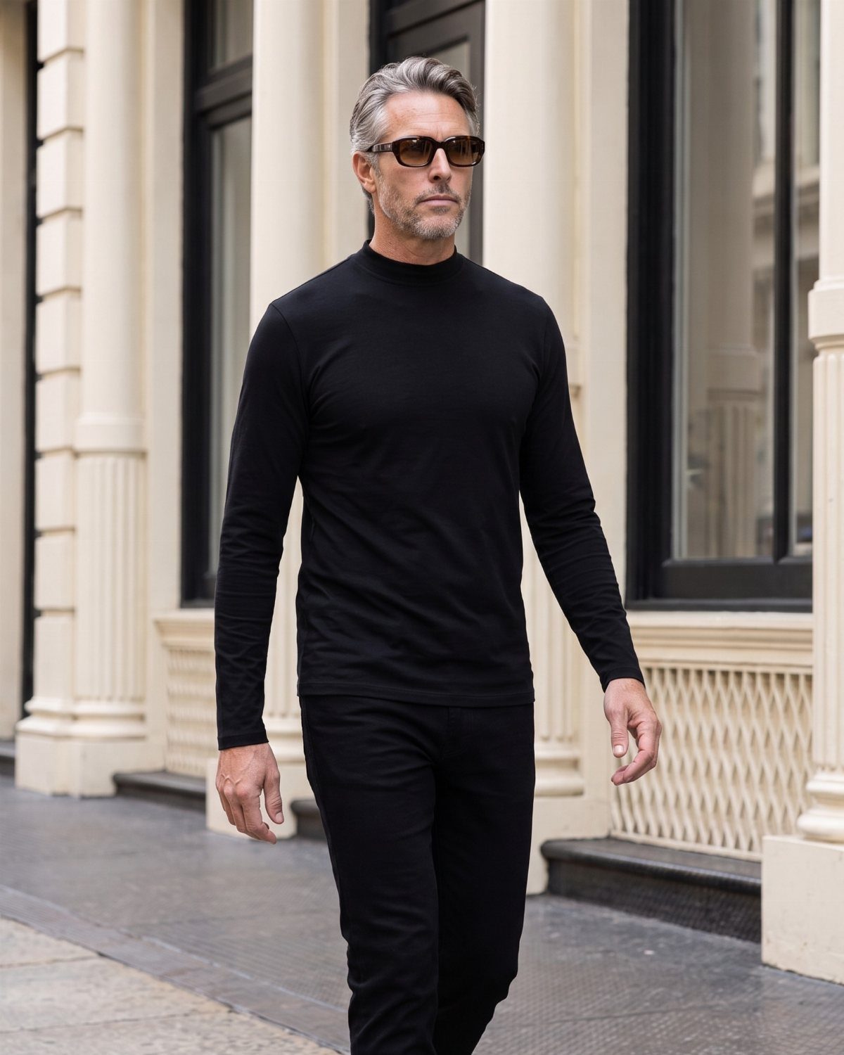 A man wearing a black mock turtleneck and black trousers walking past white columns.
