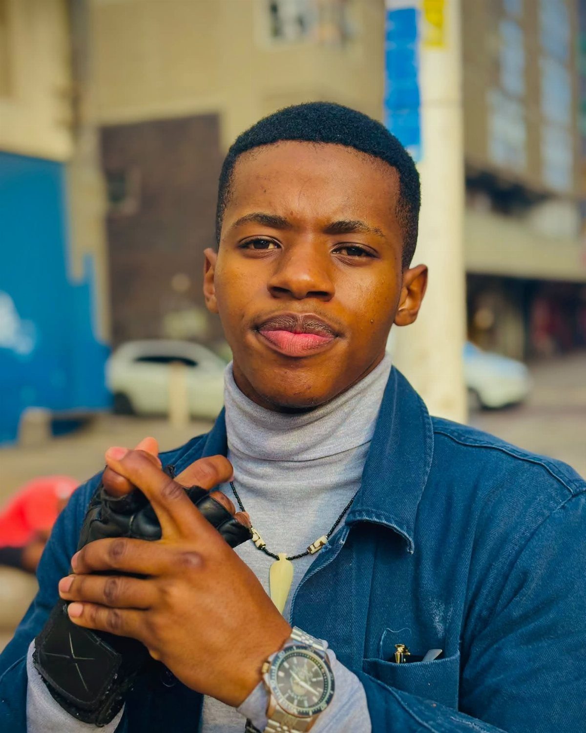 A young man wearing a grey turtleneck and a blue denim jacket while holding a camera.