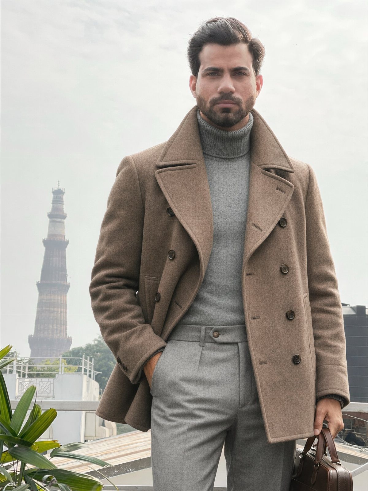 Man wearing a grey turtleneck sweater and a brown wool overcoat standing outdoors.