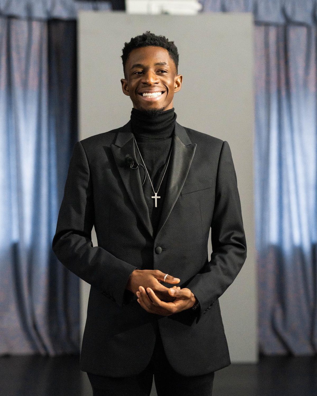 Smiling man wearing a black turtleneck sweater and a black blazer with a silver cross necklace.