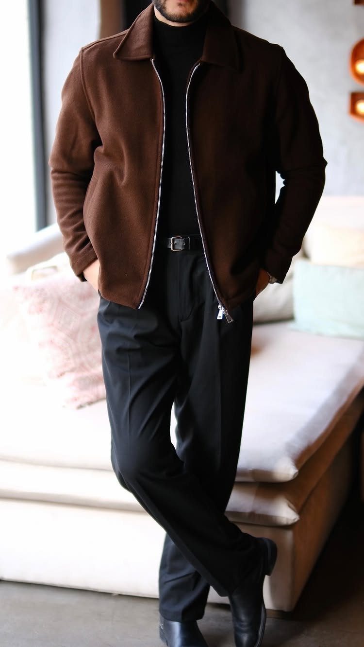 Man wearing a black turtleneck, brown jacket, and black trousers standing by a sofa.