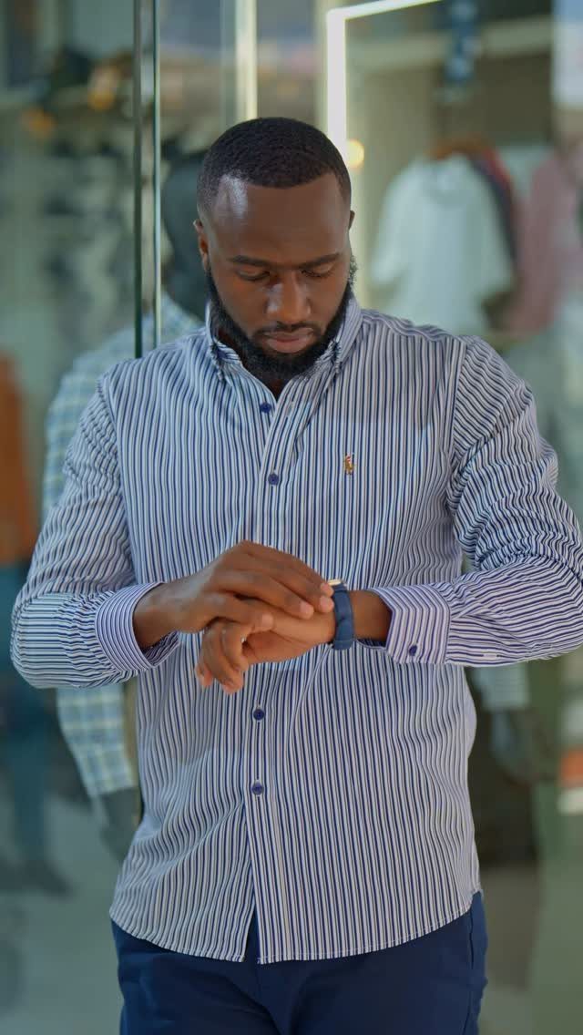 A man in a striped button-down shirt and blue trousers adjusting his wristwatch.
