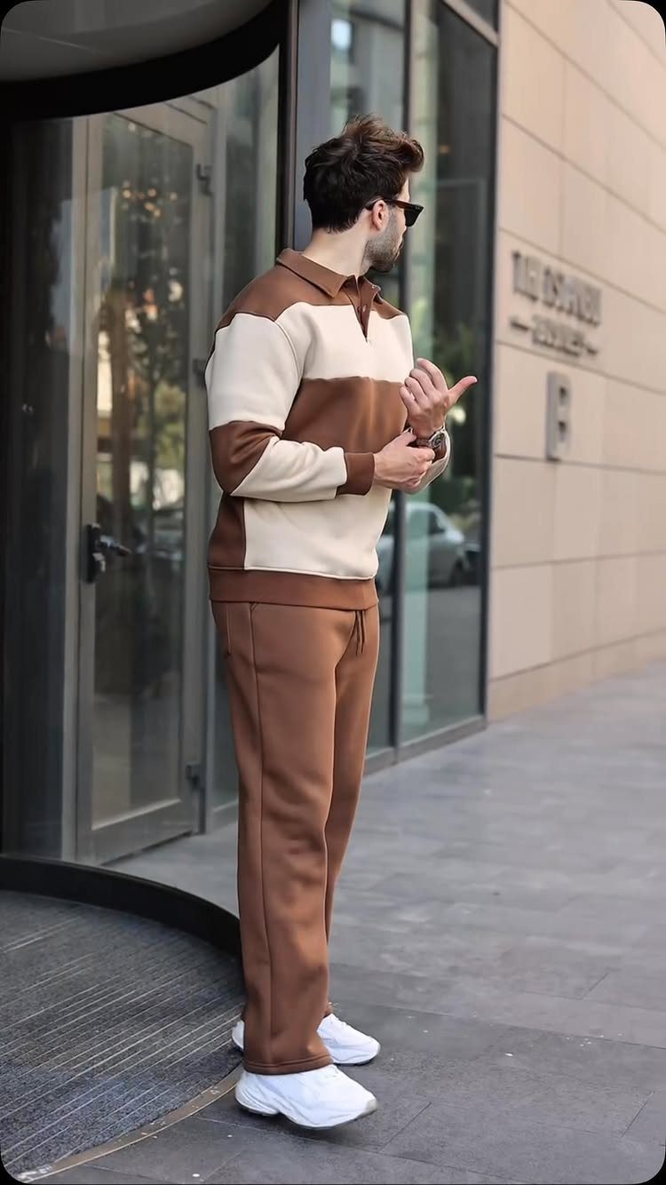 Man in a brown and beige color-blocked tracksuit and white sneakers standing by a glass door.